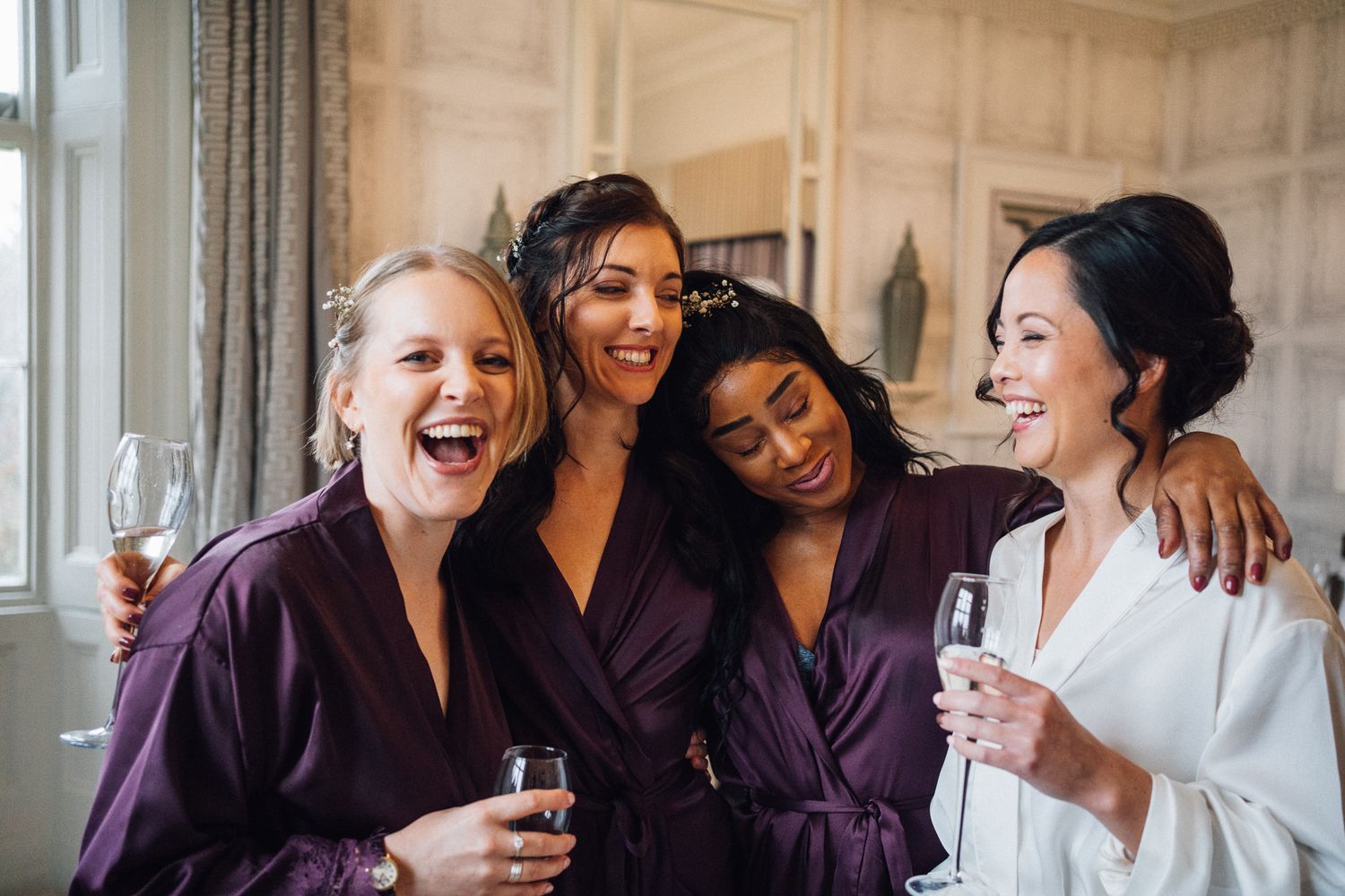 Four bridesmaids in satin robes laughing and holding champagne glasses during wedding morning preparations.
