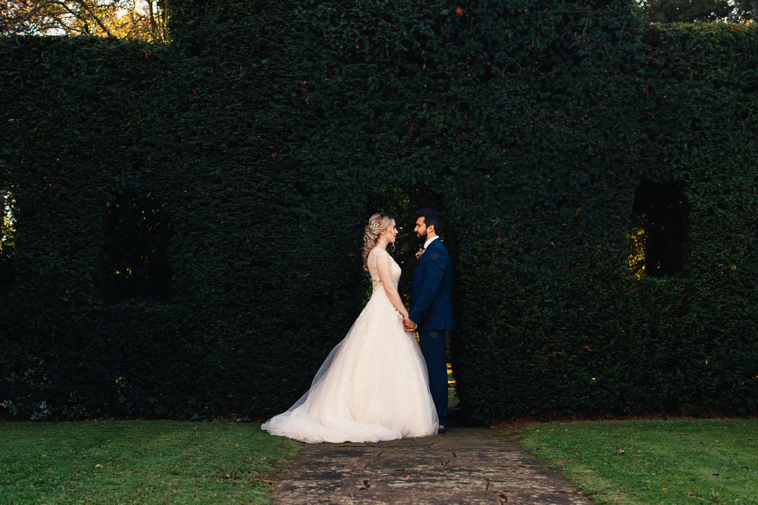 Silhouetted wedding couple poses against dark hedges in evening garden setting.