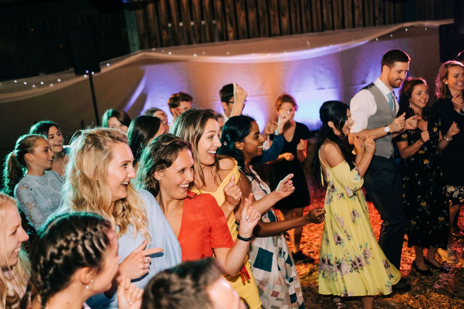 Crowd of wedding guests dancing enthusiastically under colorful party lights.