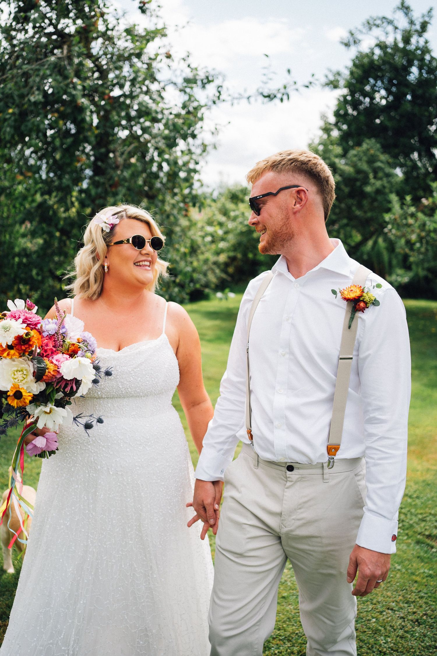 A couple in white wedding attire pose together outdoors holding colorful bouquets on a sunny summer day.