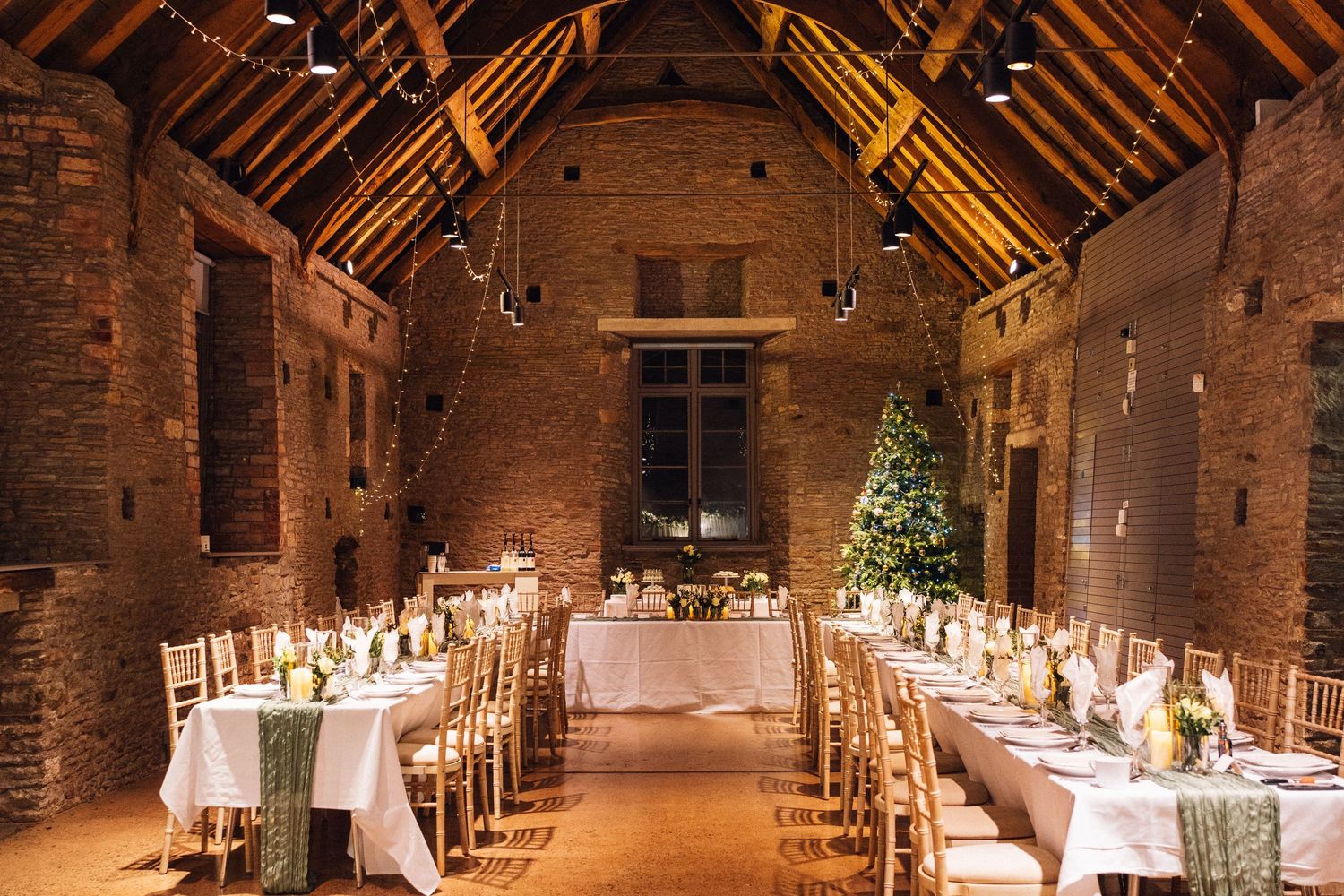 Long banquet tables set up in a rustic brick barn with wooden beams and ambient lighting.