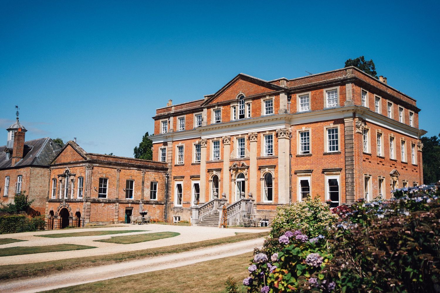 Historic red brick manor house with multiple buildings and landscaped gardens under blue sky.