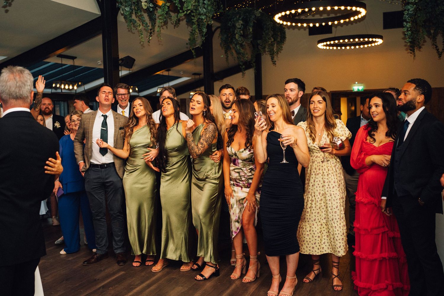 Wedding guests gather on a dance floor under circular lighting fixtures during an evening reception celebration.