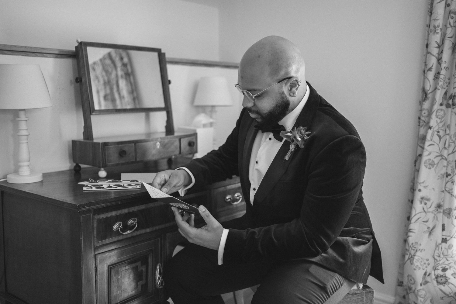 A person in a dark suit adjusts a boutonniere while getting ready in a room with floral curtains and lamps.