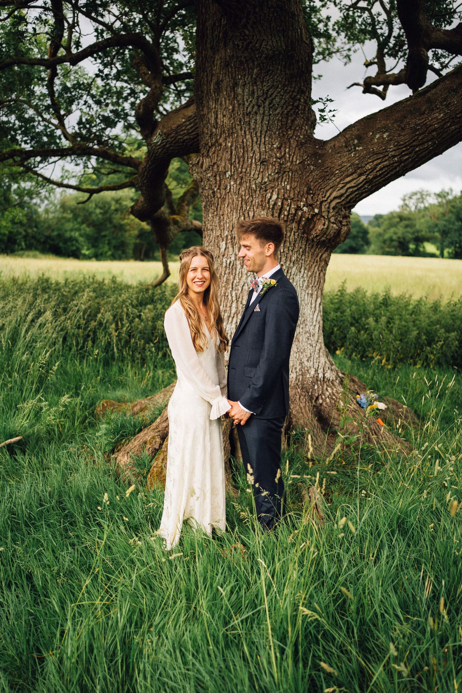 Wedding couple poses in front of a large tree in a lush green field during golden hour.