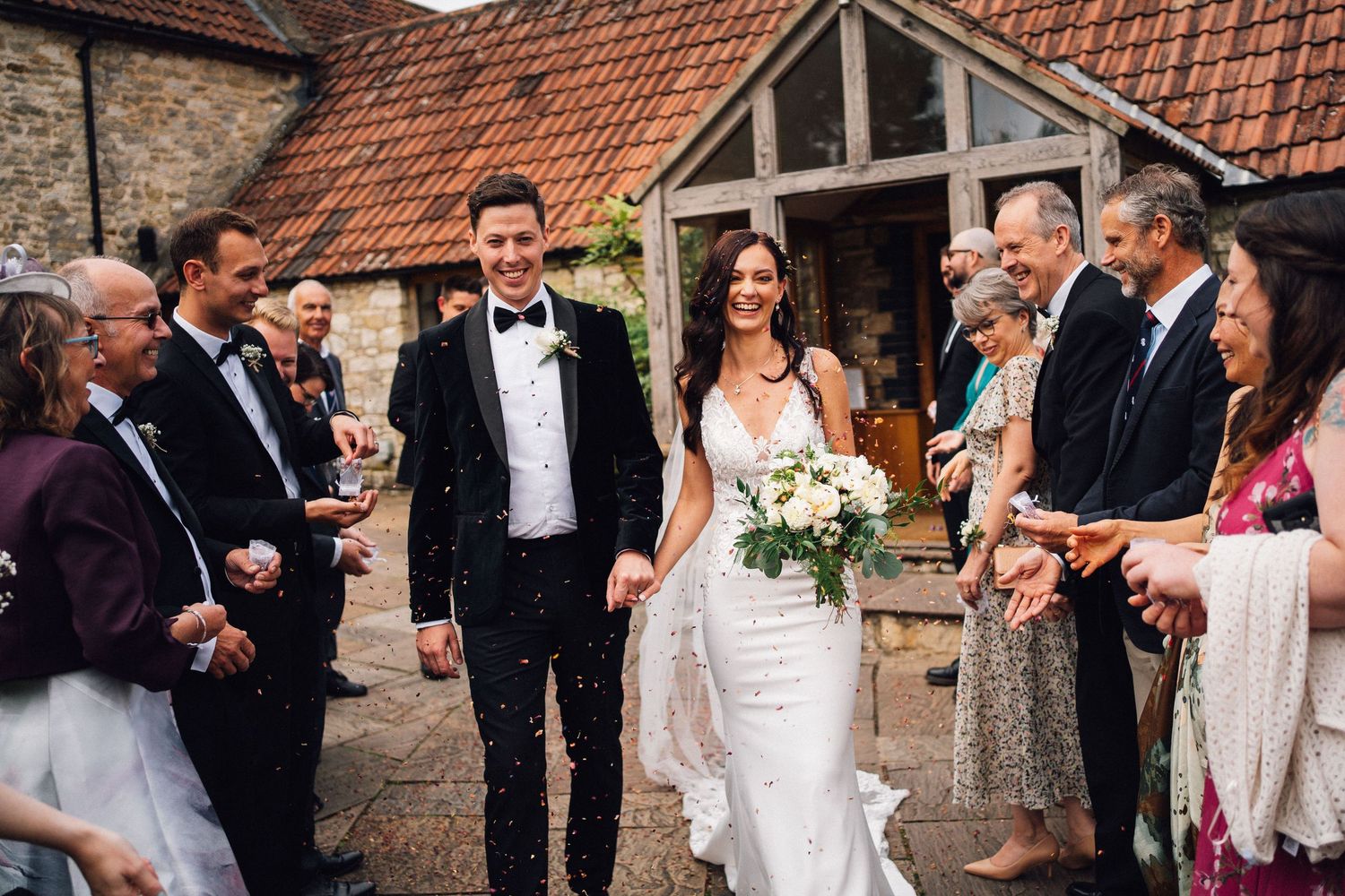 Wedding guests gather outside a rustic stone building with red tile roof during a celebration.