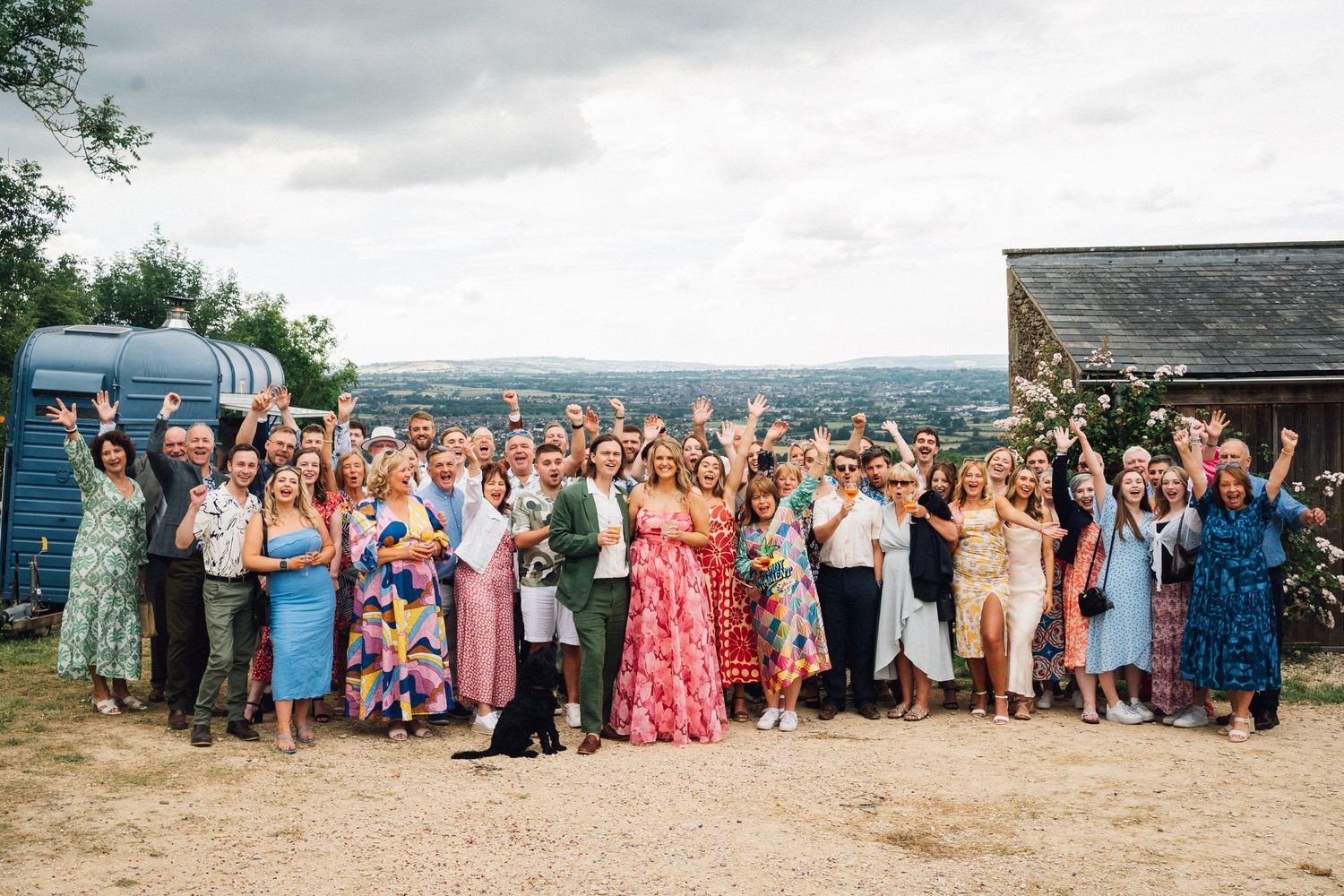 Wedding party and guests gather for a group photo against a scenic countryside backdrop.