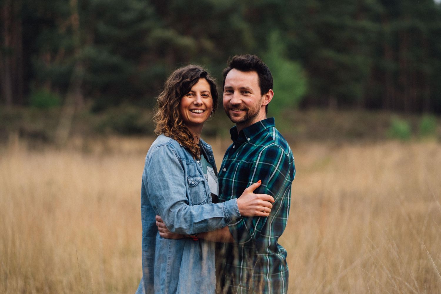 A couple laughs together in a golden field with evergreen trees in the background.