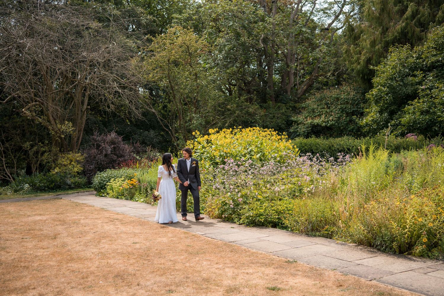 Couple in wedding attire standing on a garden path surrounded by lush greenery and colorful flowers.