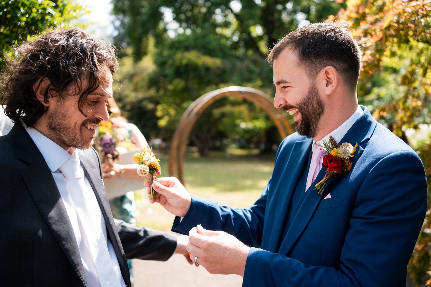 Two grooms share a joyful moment pinning boutonnieres at an outdoor wedding ceremony.