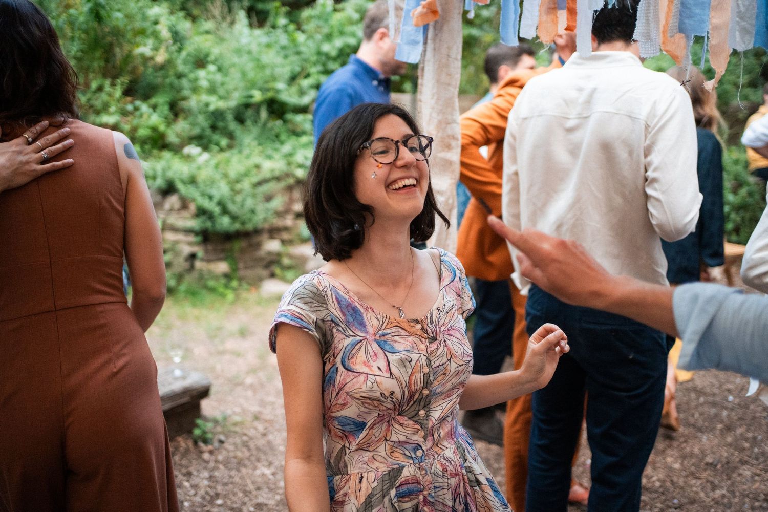 A woman in a floral dress laughs joyfully at an outdoor wedding reception surrounded by guests.