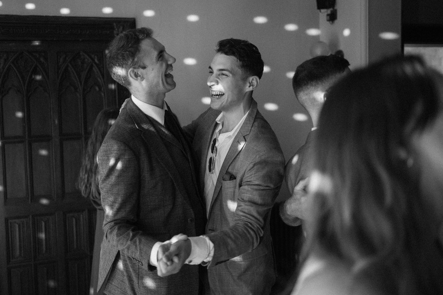 Two men in suits share a joyful handshake and smile at a wedding reception in black and white.