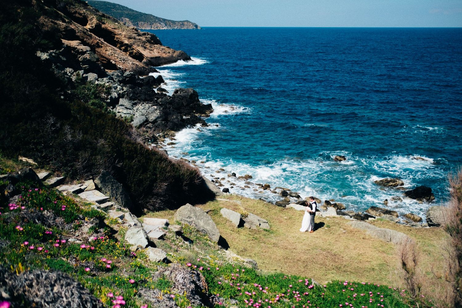 Rocky coastline with turquoise ocean waves crashing against cliffs under bright blue sky.