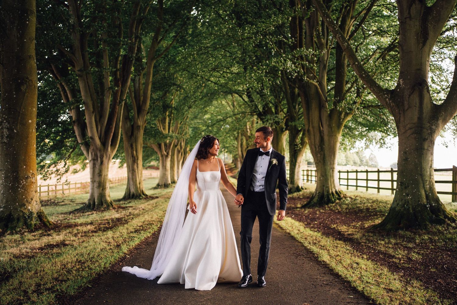 Newlyweds walking hand in hand down tree-lined path in elegant wedding attire.