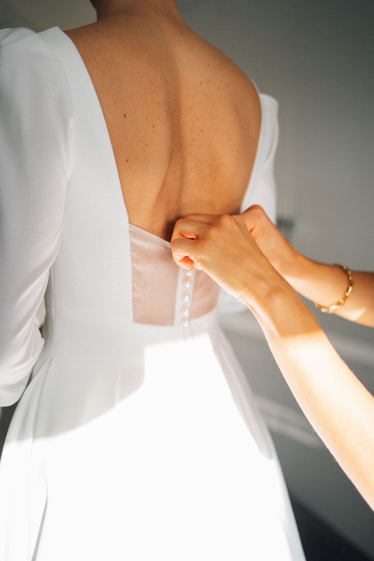 Hands fastening buttons on the back of an elegant white wedding dress in soft natural lighting.