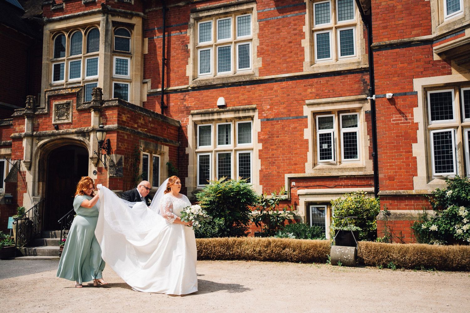 Bride in white gown with bridesmaids adjusting her train outside a stunning red brick Victorian wedding venue.