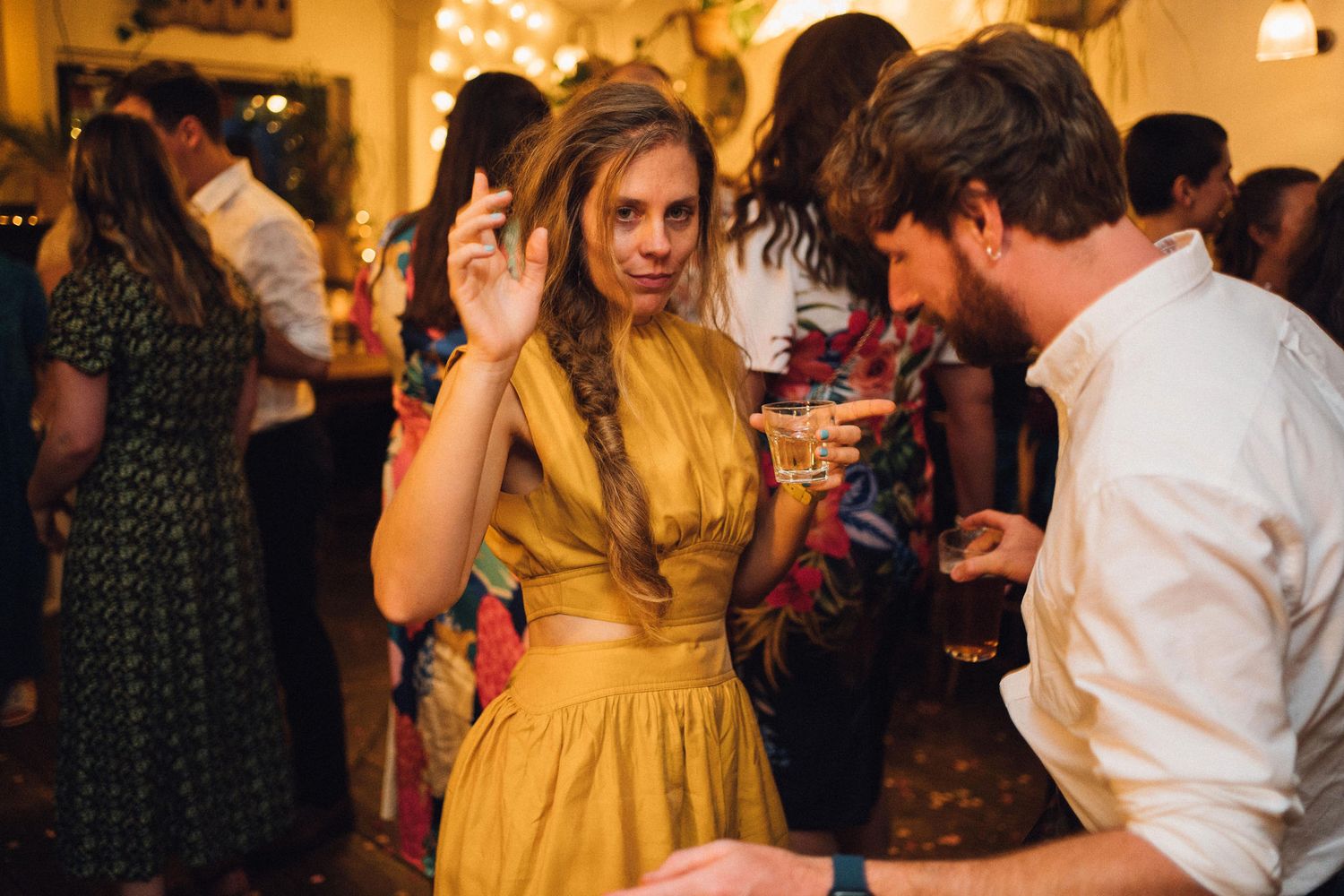 A person in a golden yellow dress gestures while talking to someone at a crowded indoor party.
