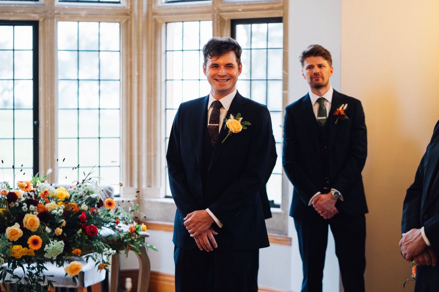 A wedding party stands in a bright room with large windows and floral arrangements, dressed in formal black attire.