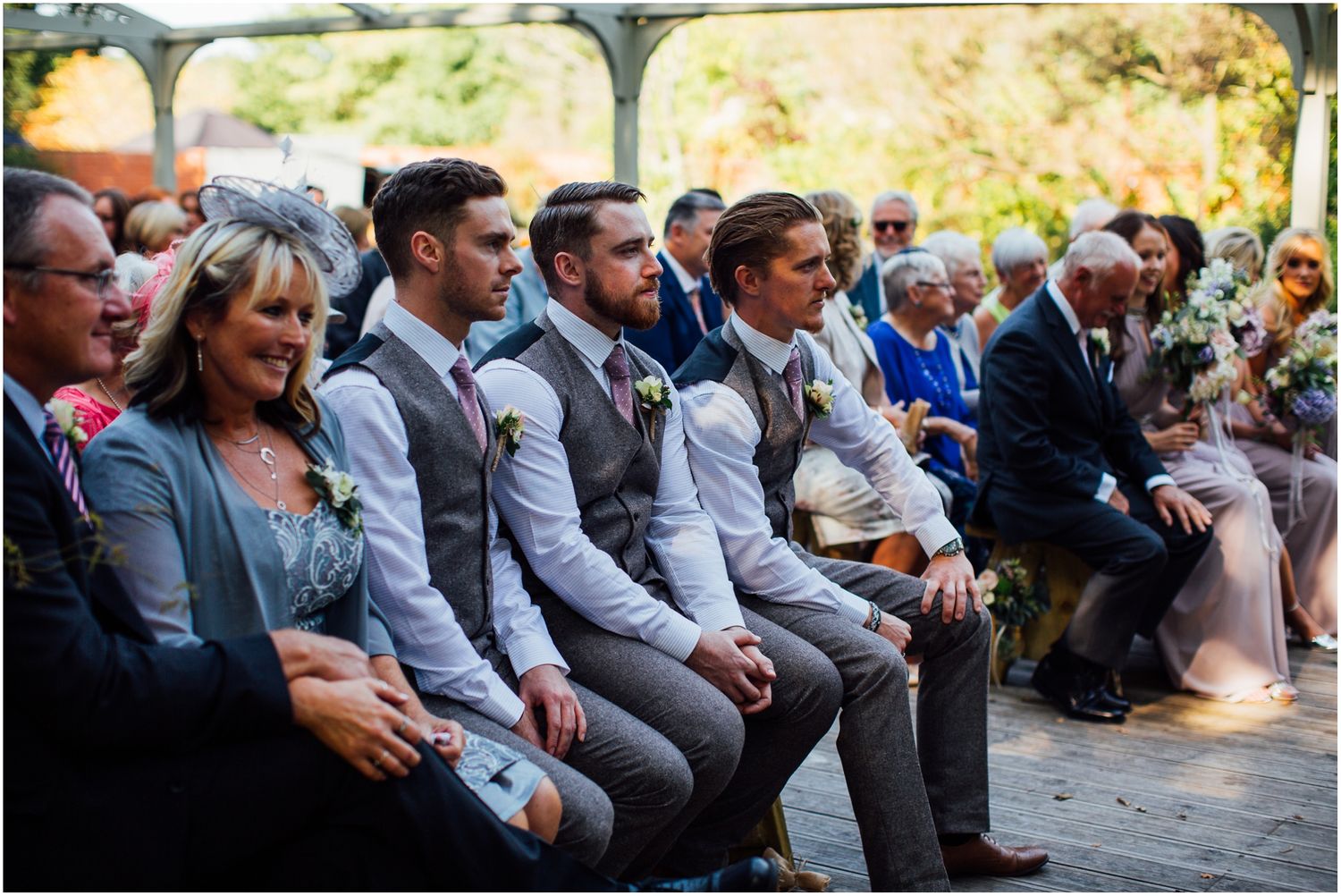 Wedding guests in grey suits seated outdoors during ceremony.