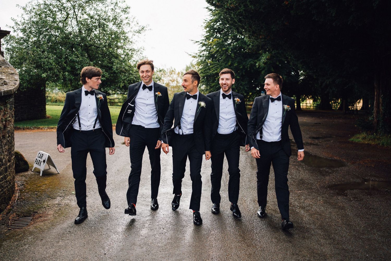 Wedding party members in black suits walking in formation on a garden pathway.
