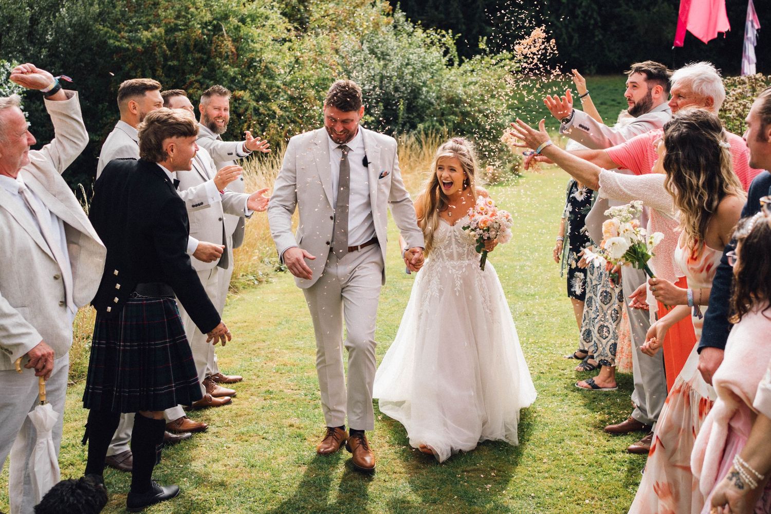 Wedding guests throw confetti as newlyweds walk down a grassy aisle during an outdoor celebration.