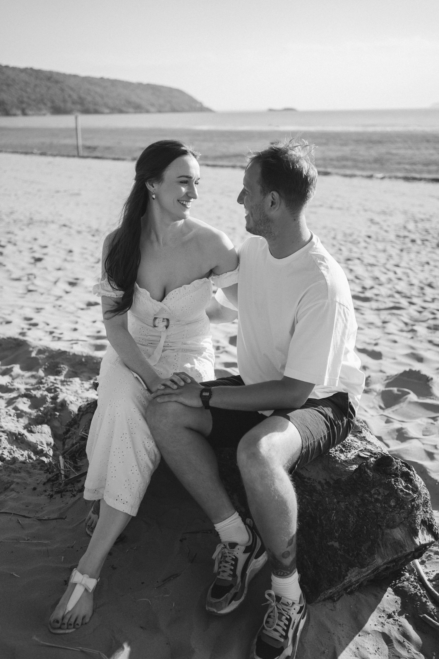 Black and white photo of couple sitting on rocky beach during engagement session.