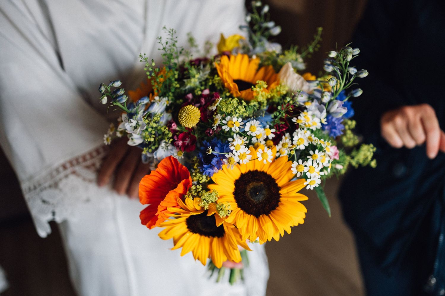 Vibrant sunflower and wildflower wedding bouquet with blue accents against white background.