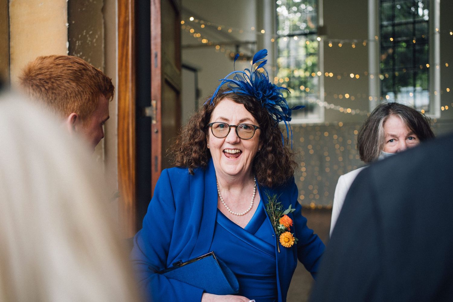 A guest in a bright blue dress and matching fascinator smiles during a celebration event with string lights in background.