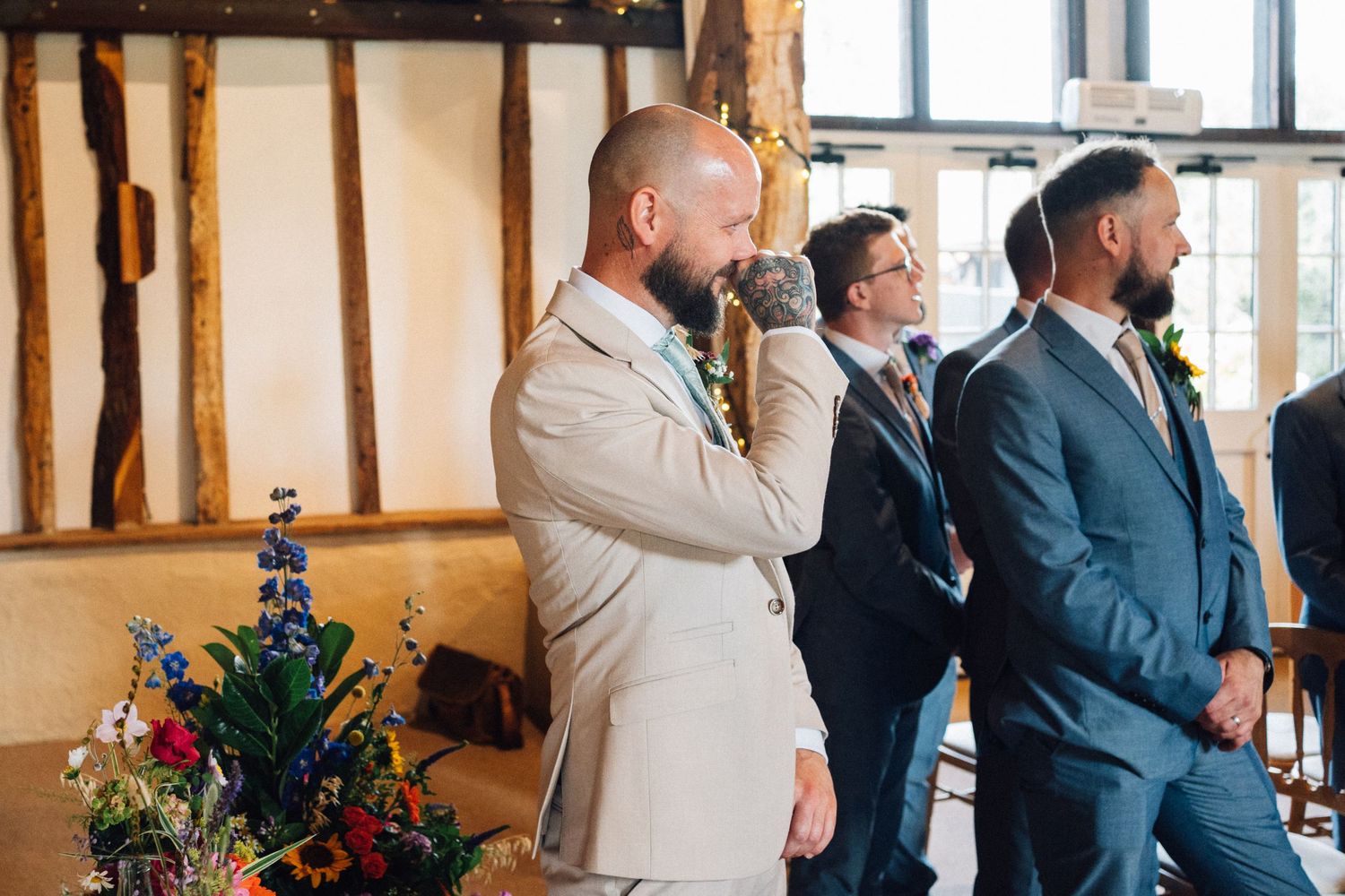 Wedding guests in formal attire stand together during a reception in a rustic venue with wooden beams and floral decor.