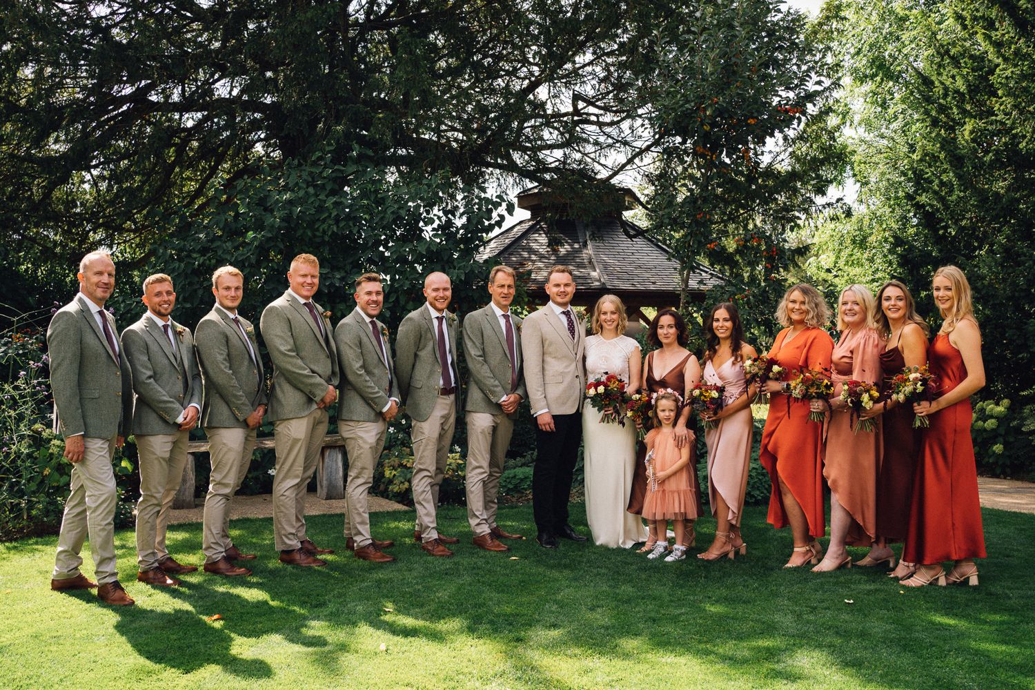 Wedding party members in gray suits and coral dresses pose together on a grassy lawn during an outdoor celebration.