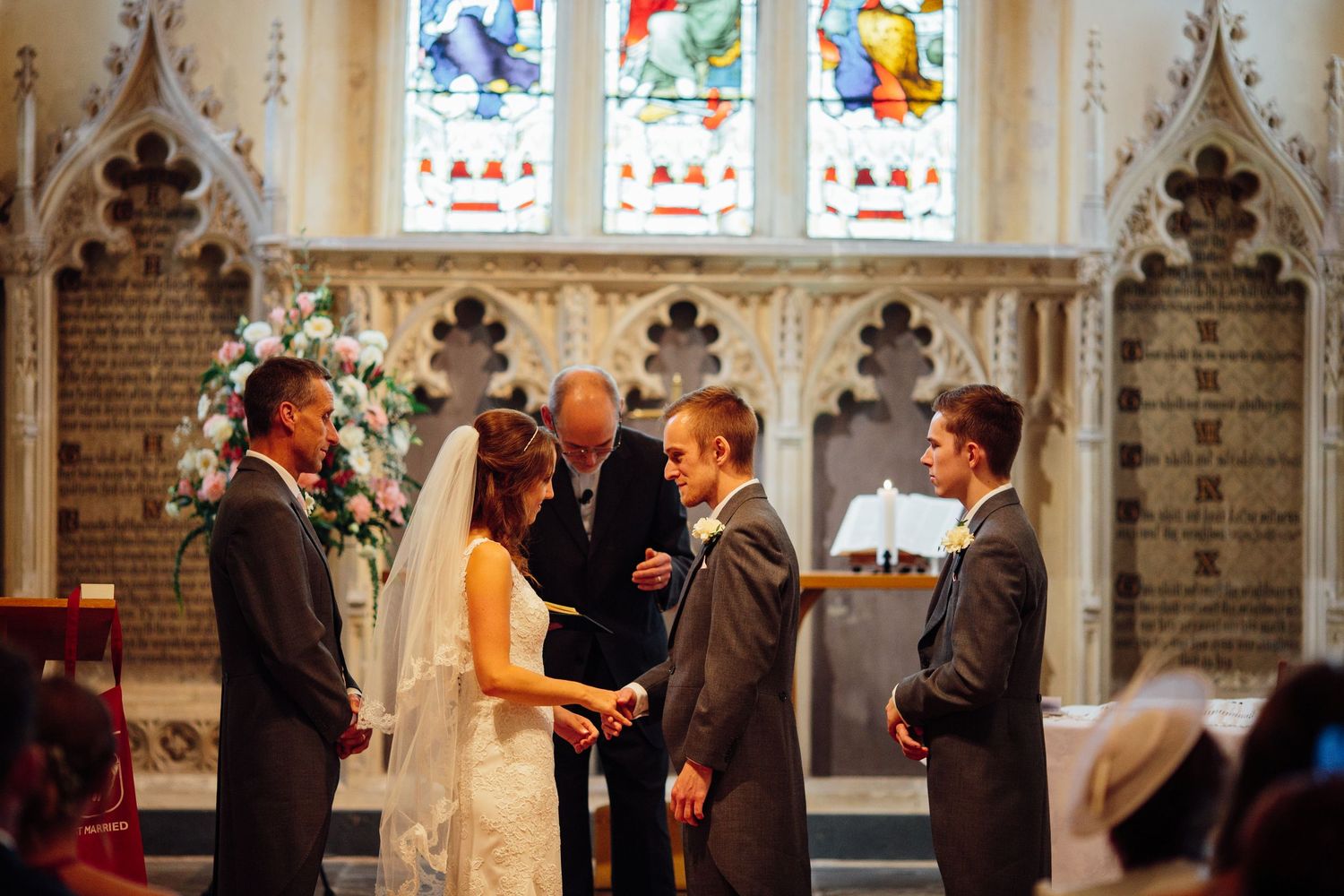 Church wedding ceremony with people gathered in front of ornate stained glass windows and stone architecture.