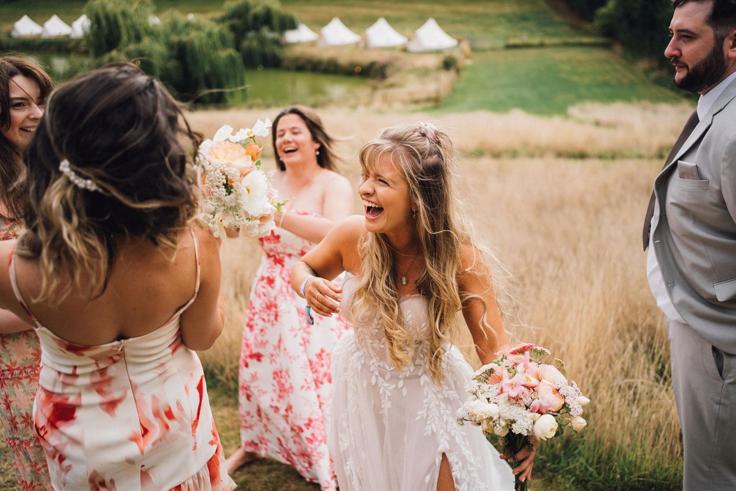 Wedding party wearing floral dresses laughs together in a sunlit field during golden hour.