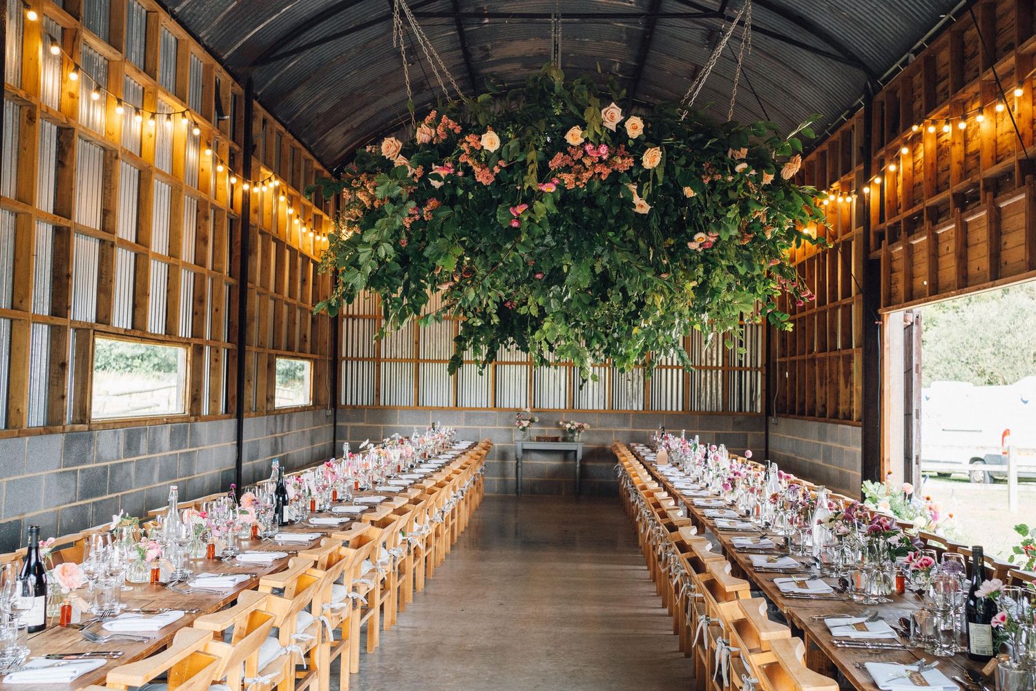 Long rustic reception tables decorated with string lights and floral arrangements in an elegant barn venue.