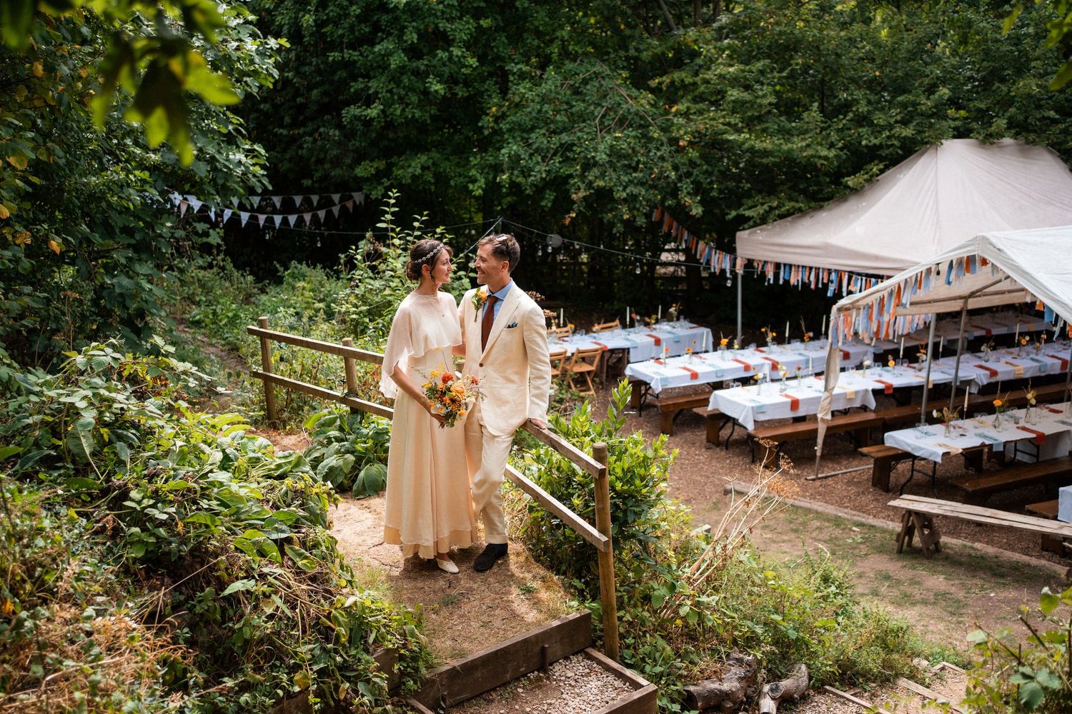 Newlywed couple stands on garden steps overlooking festive outdoor wedding reception with long decorated tables.