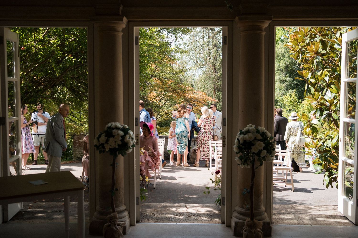 Elegant wedding ceremony viewed through grand open doors, with guests gathering on a sunlit garden pathway.