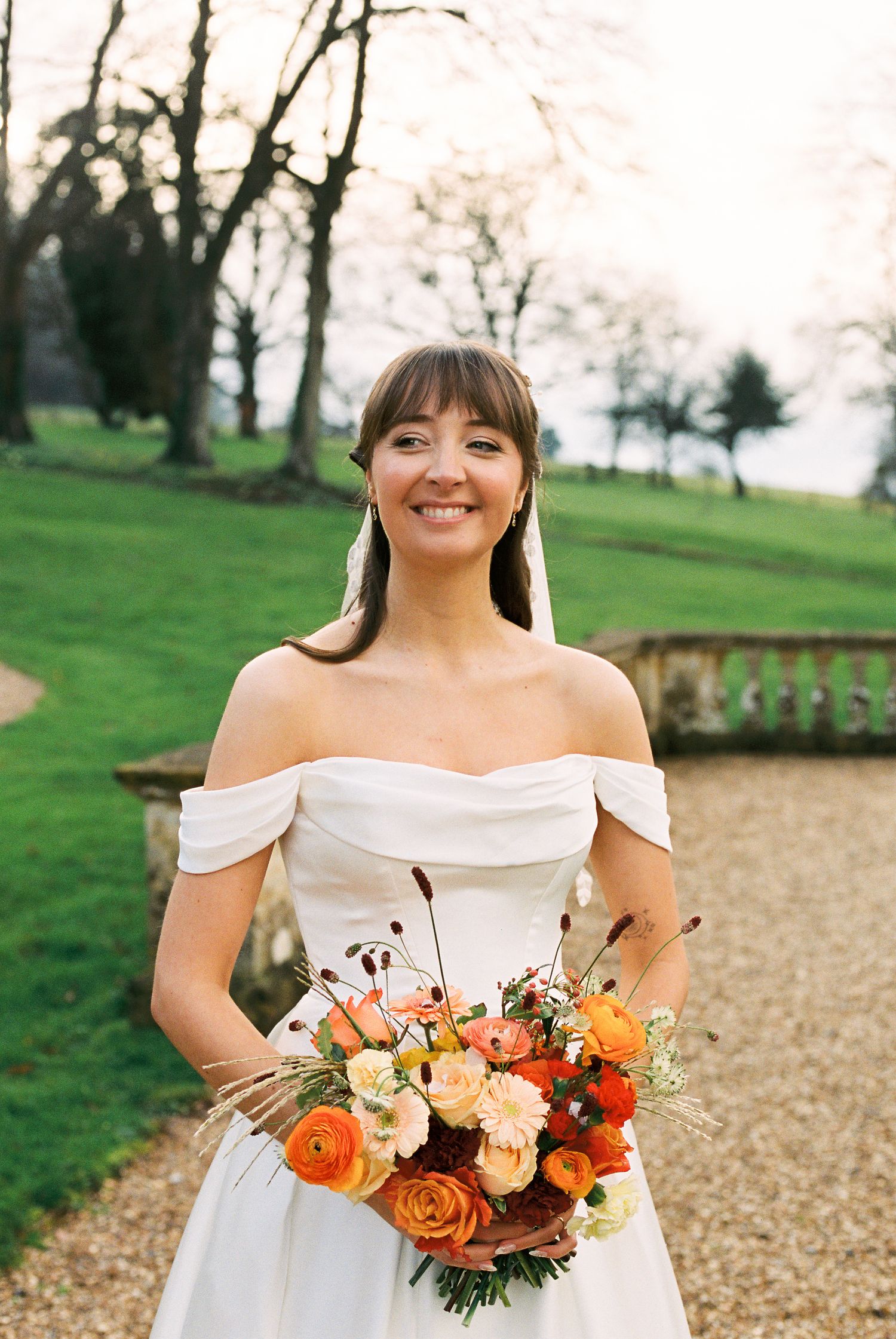 Smiling bride in off-shoulder white gown holding vibrant orange and peach bouquet, standing outdoors on gravel path.