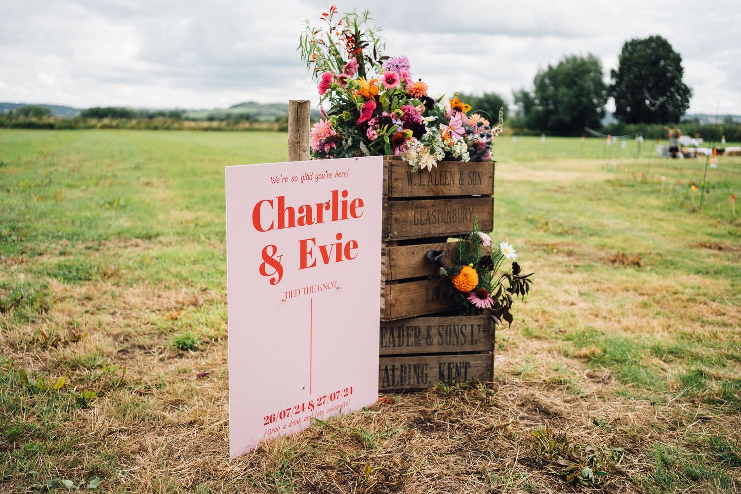 Rustic wooden crates with colorful floral arrangements decorate an outdoor field setting for a wedding ceremony.