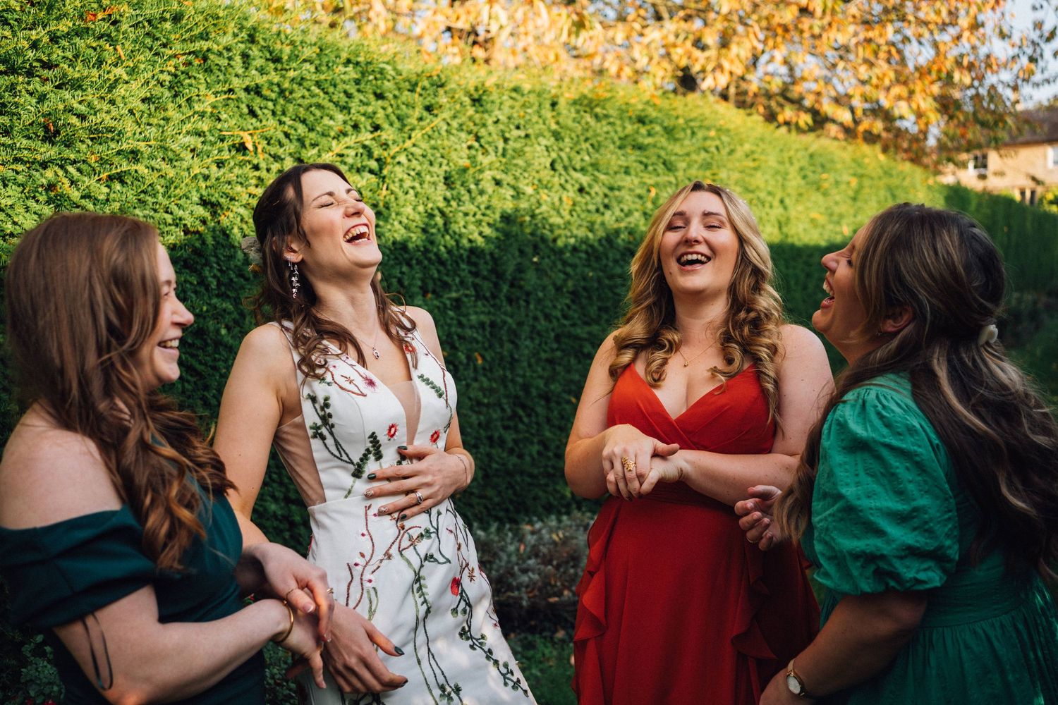Four women in formal dresses share a candid laughing moment in a garden setting.