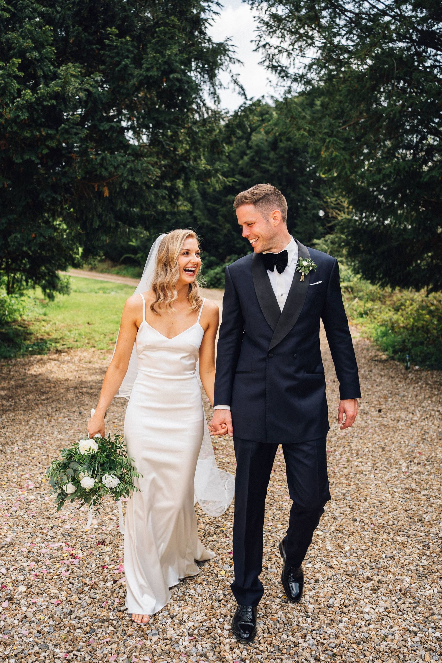 Couple walking on gravel path surrounded by trees in formal attire.