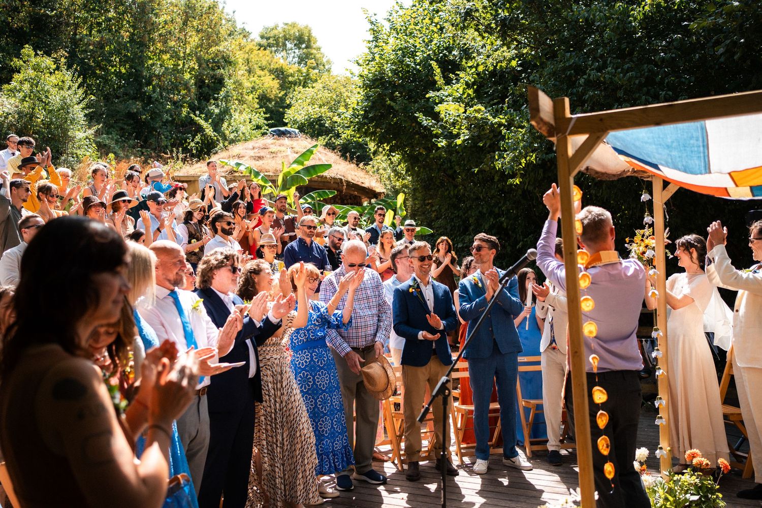 Joyful wedding ceremony outdoors with guests applauding under sunny skies near a wooden arch.