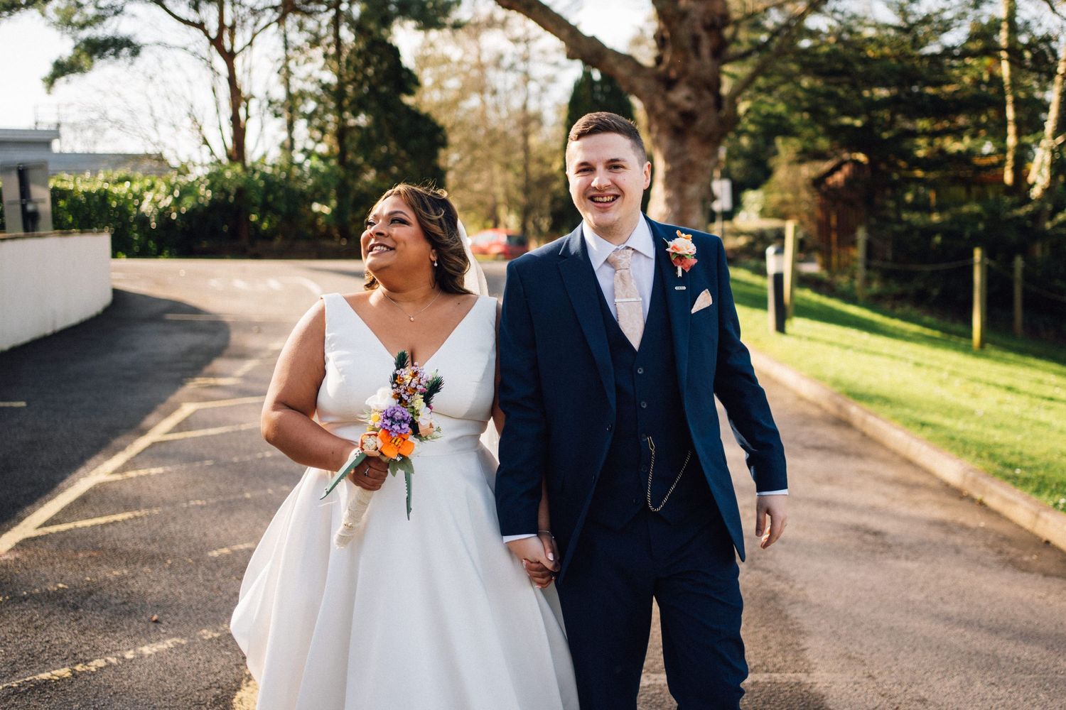 A couple in wedding attire walks down a tree-lined path during golden hour, with the bride wearing a flowing white gown.