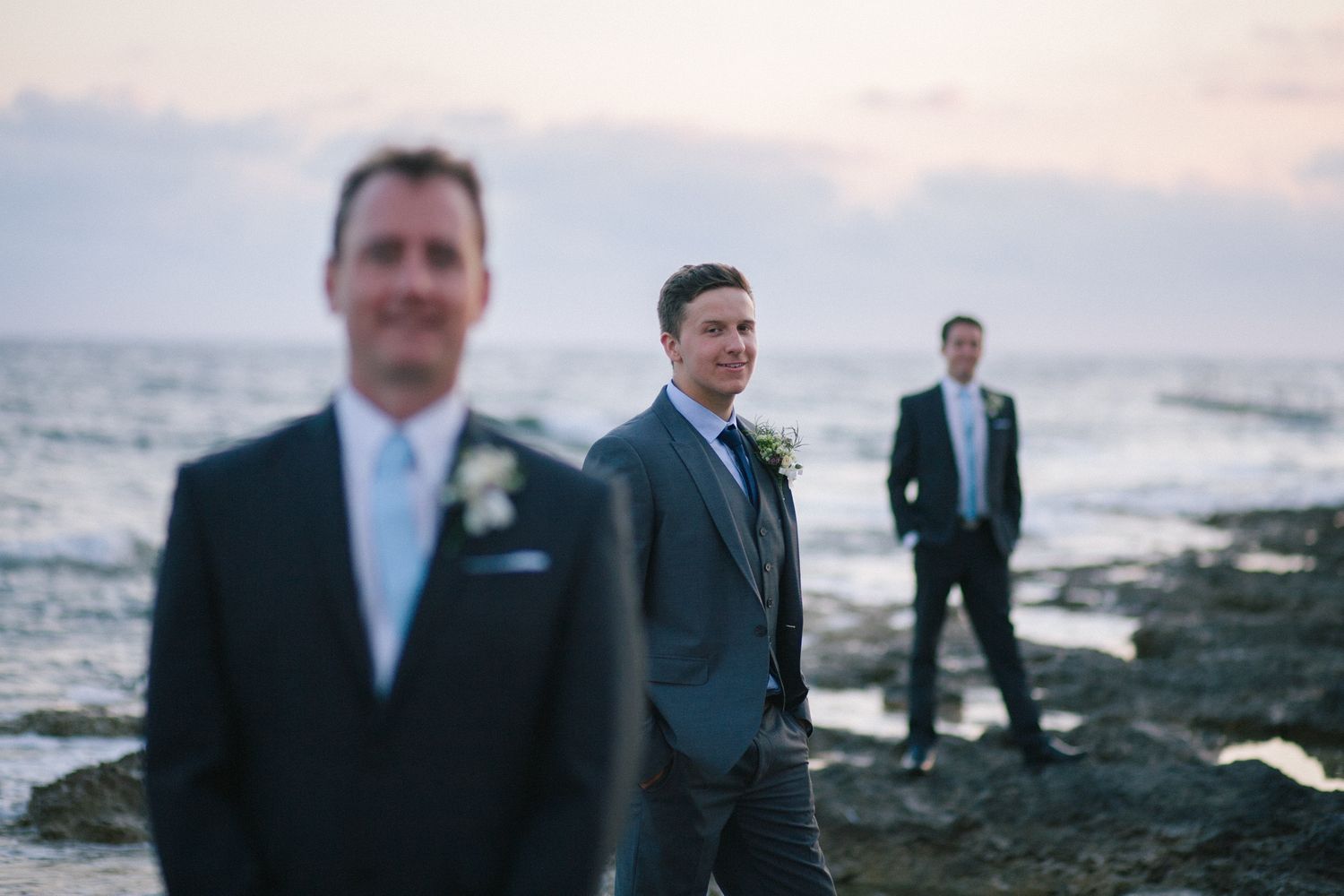 Wedding party members stand on a rocky beach at sunset.