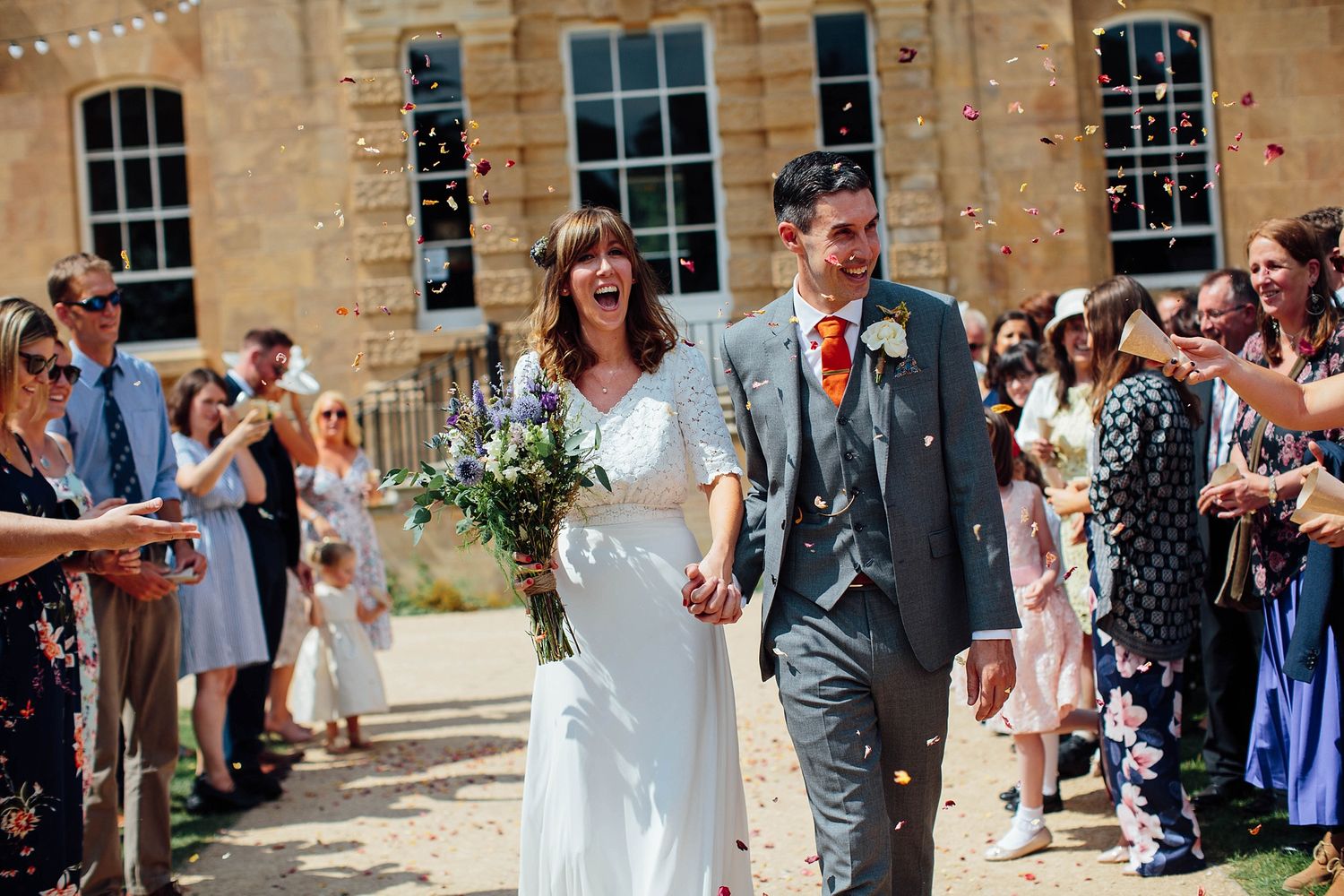Newlyweds walk through confetti shower outside historic stone building during wedding celebration.