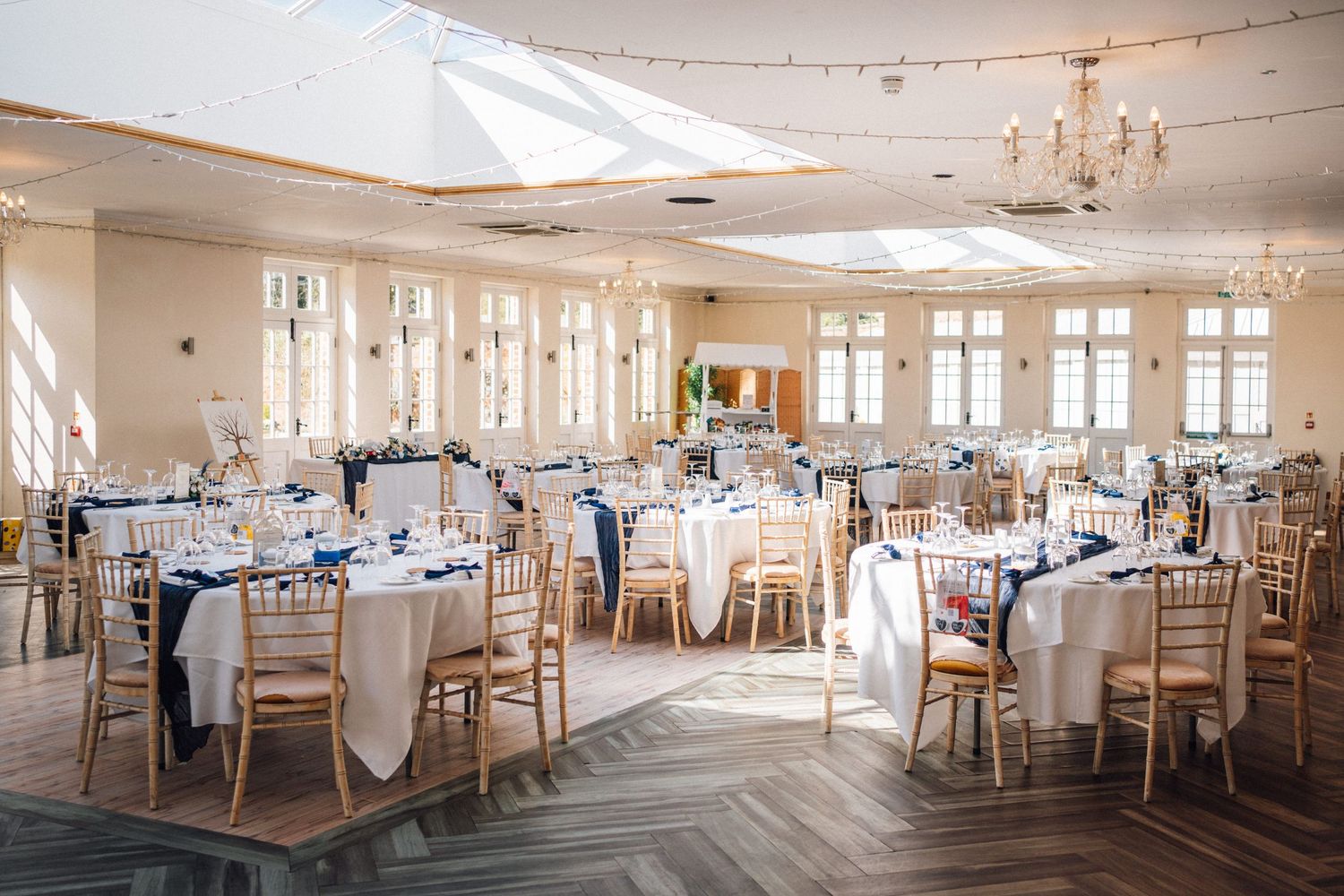 Elegant wedding reception hall with round tables, white linens, navy accents, gold chiavari chairs, and natural light from large windows.