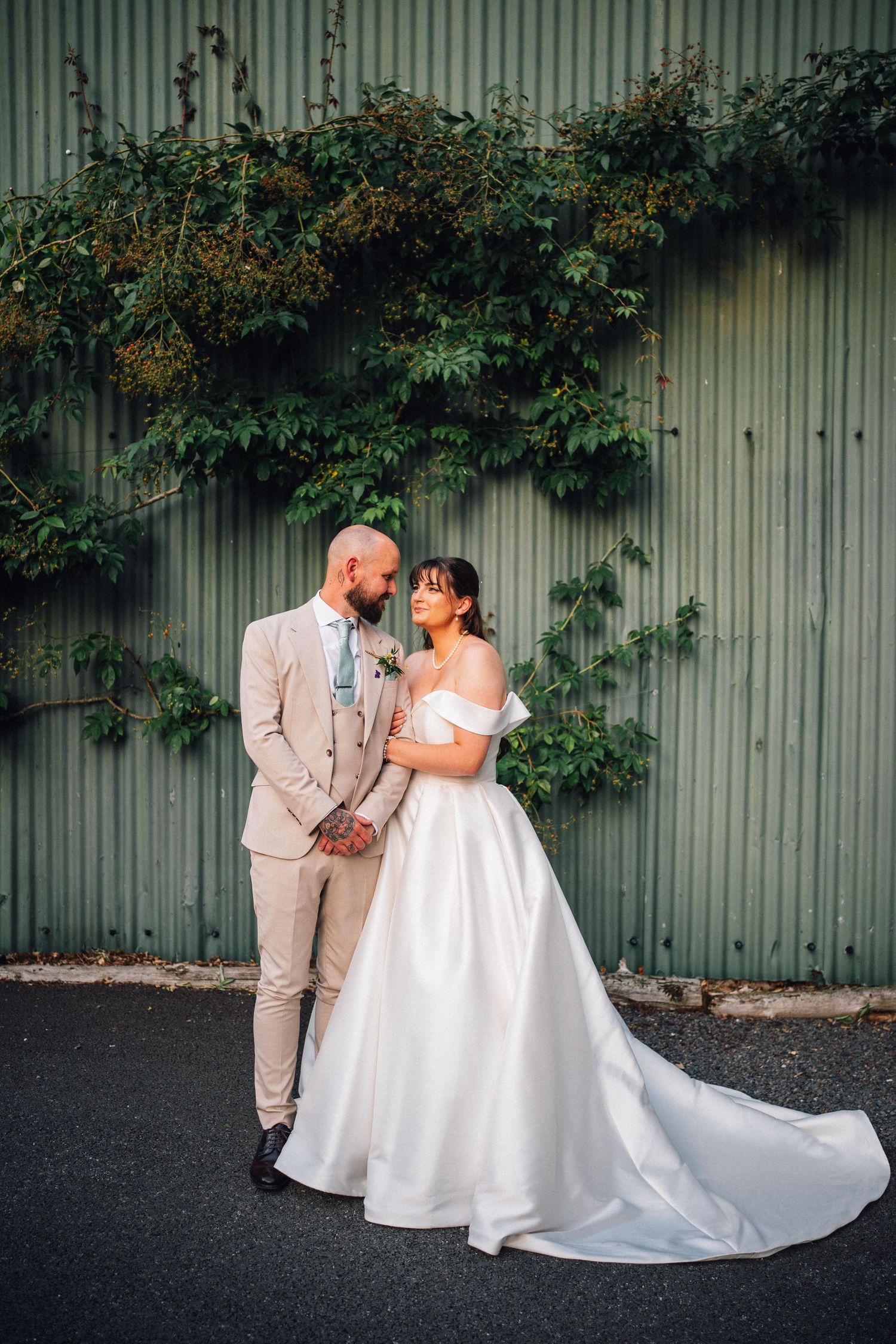 Bride in white off-shoulder gown and groom in beige suit hold hands before a green ivy-covered wall.