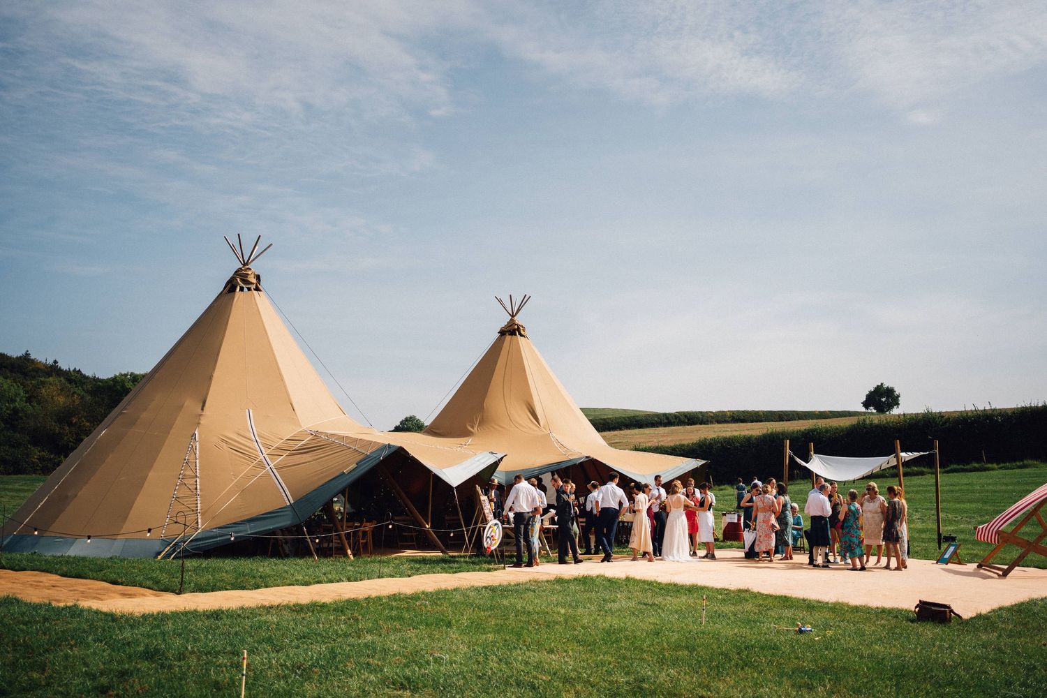 Outdoor wedding reception with two large tipi tents on a green field under a blue sky.