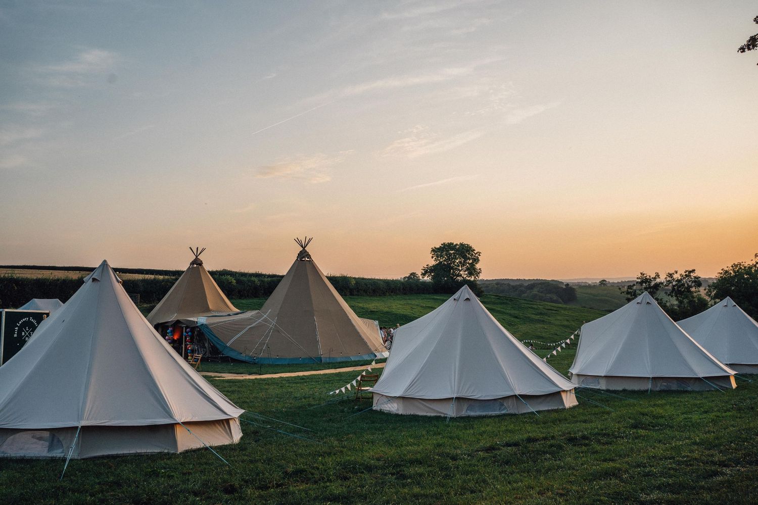White bell tents and tipis arranged on green grass at sunset during a glamping festival event.