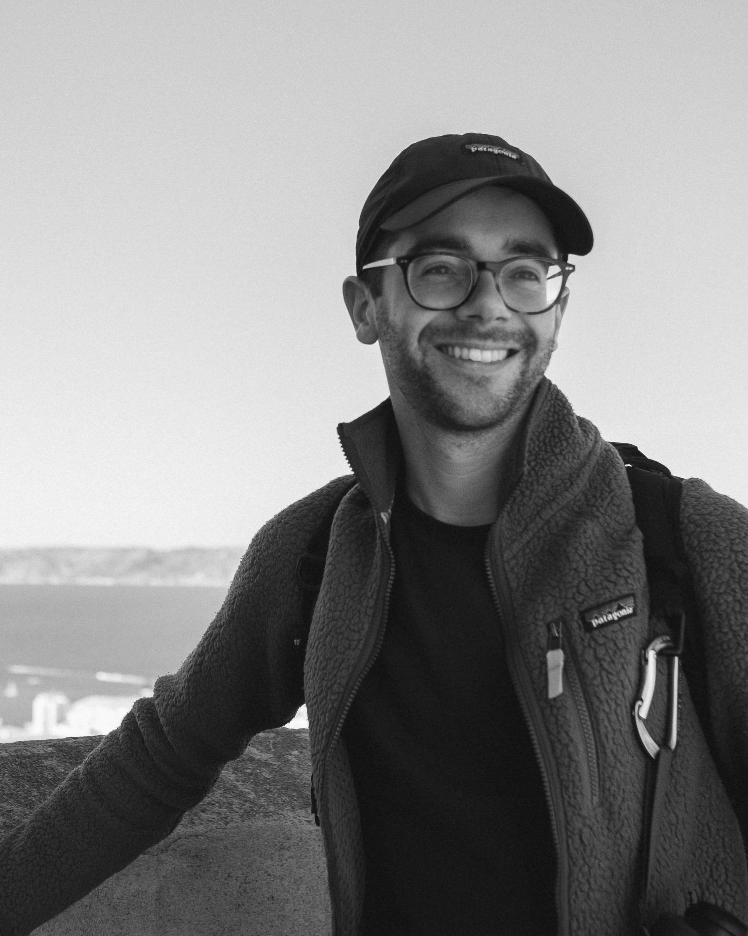 Smiling young man wearing glasses and a cap outdoors with a scenic coastal view in black and white.