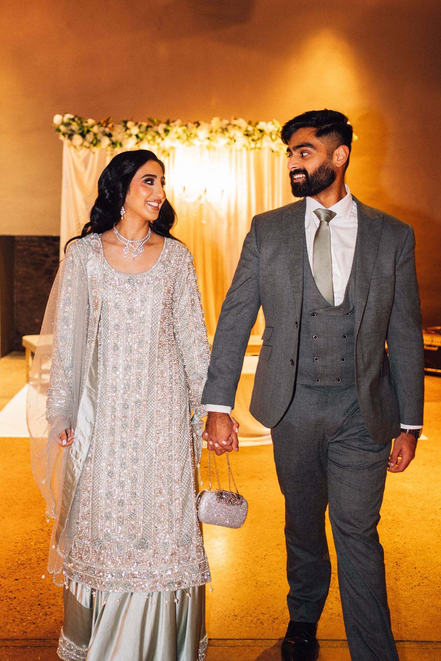 A couple in formal attire walks together, with the bride wearing a silver embellished dress and groom in a gray suit.