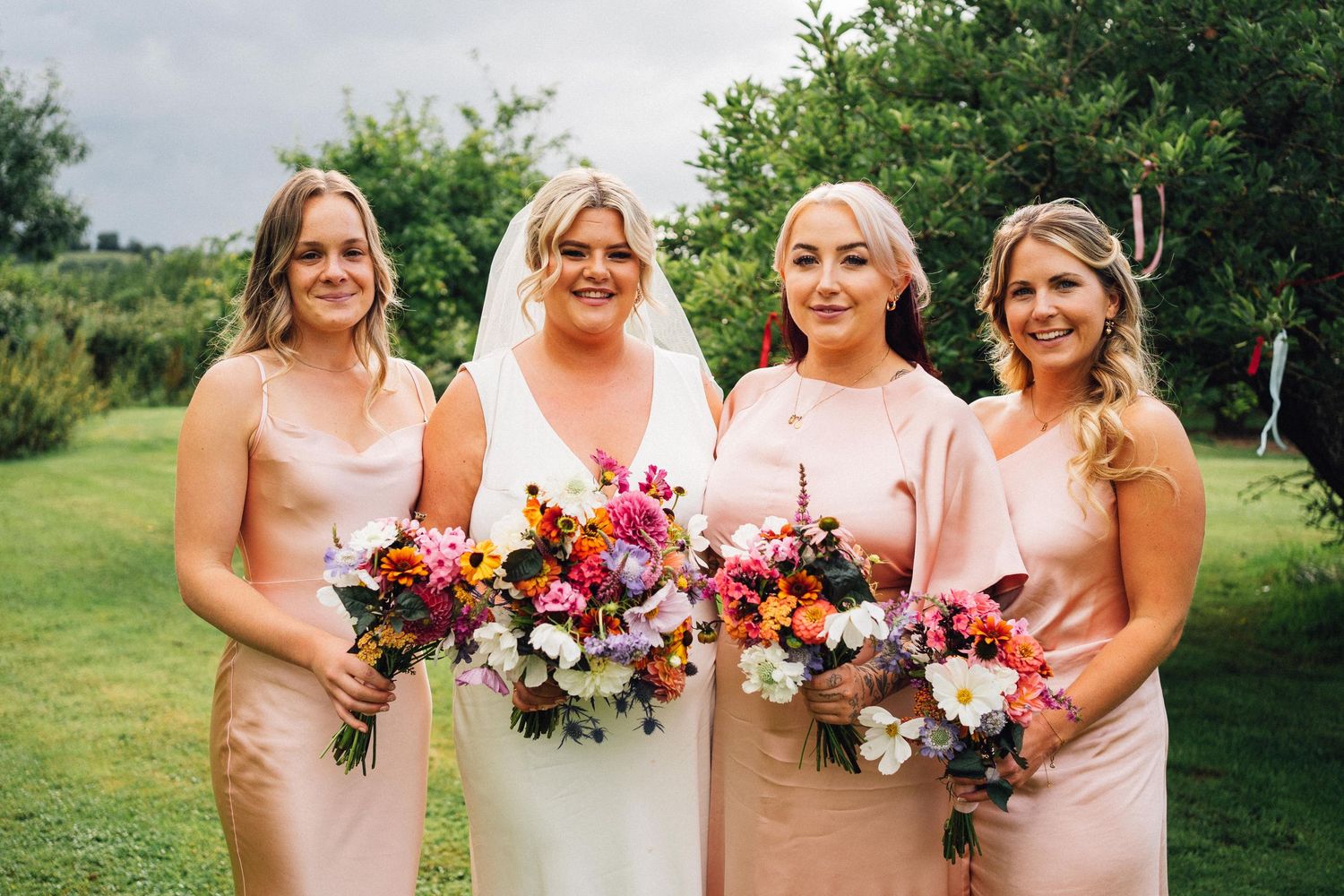 A group of bridesmaids in pink dresses pose together holding vibrant floral bouquets in an outdoor garden setting.